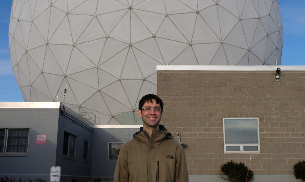 A man standing in front of the Haystack 37m radio antenna's white radome enclosure