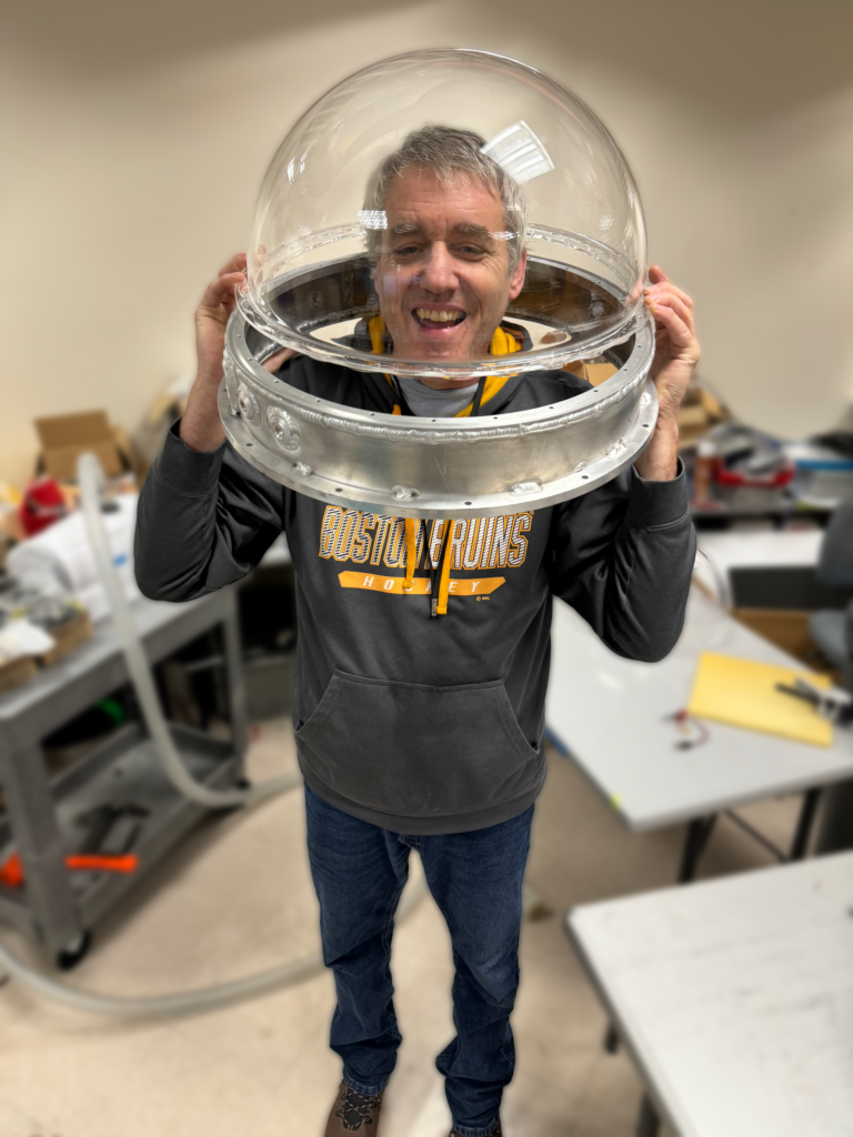 A man standing and grinning with a clear dome and metal collar over his head